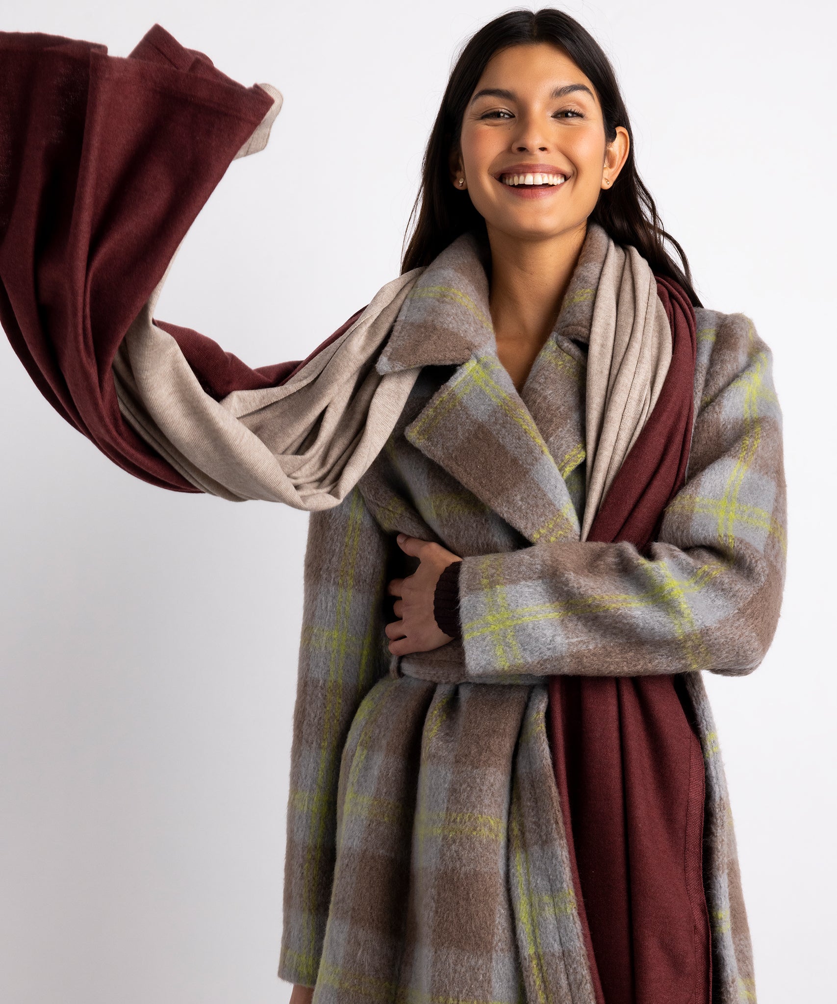 Model wearing two Essential Travel Wraps around her neck.  One is the brick colorway and one is the farro colorway.  She is wearing a plaid coat and smiling.  One side of the wraps has been thrown in the air.