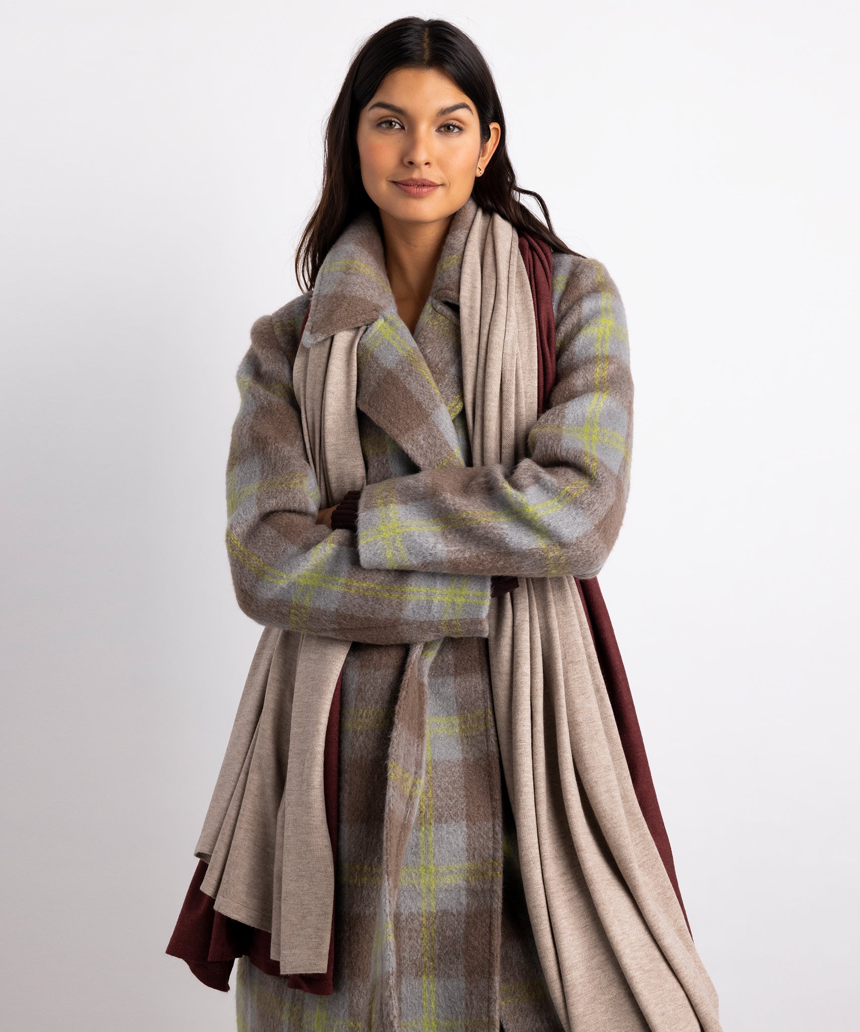 Model wearing two Essential Travel Wraps around her neck.  One is the brick colorway and one is the farro colorway.  She is wearing a plaid coat..